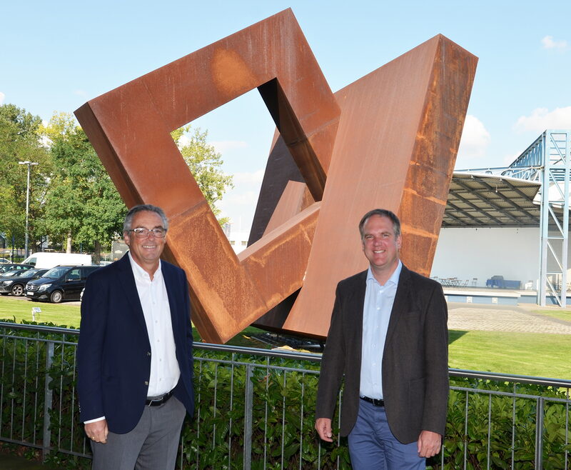 Bürgermeister Dirk Breuer und Professor Wolfgang Prinz vom Fraunhofer FIT stehen vor einer Skulptur auf dem Gelände der ehemaligen MMC-Studios.