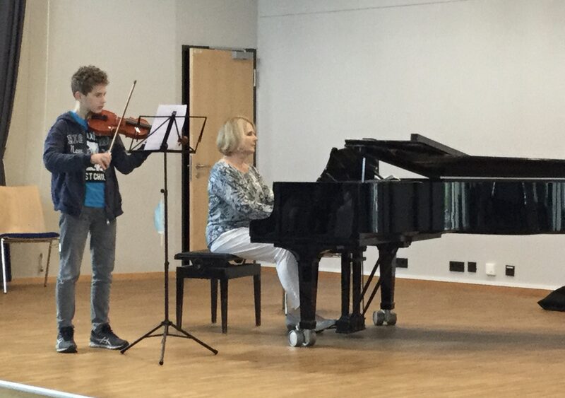 Kind steht vor Notenständer mit einer Geige. Neben ihm begleitet eine Frau auf einem Klavier.