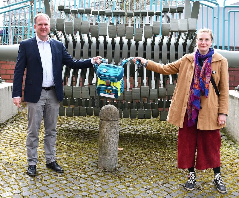 Julia Fehlisch und Bürgermeister Dirk Breuer präsentierten einen Schulrucksack vor einem stillgelegten Läufer einer Kohlenmühle zwischen Rat- und Bürgerhaus.