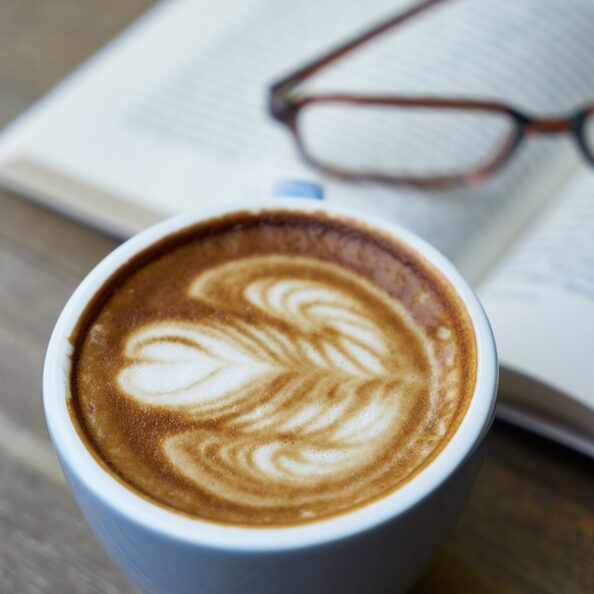 Cappuccinotasse mit Herz im Schaum im Vordergrund, aufgeschlagenes Buch und Brille im Hintergrund